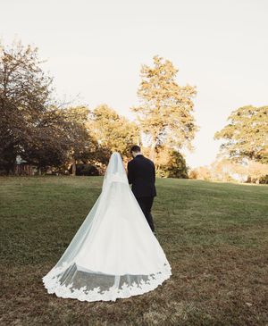 Bride and groom walk across green lawn at sunset, cathedral lace veil trailing behind.