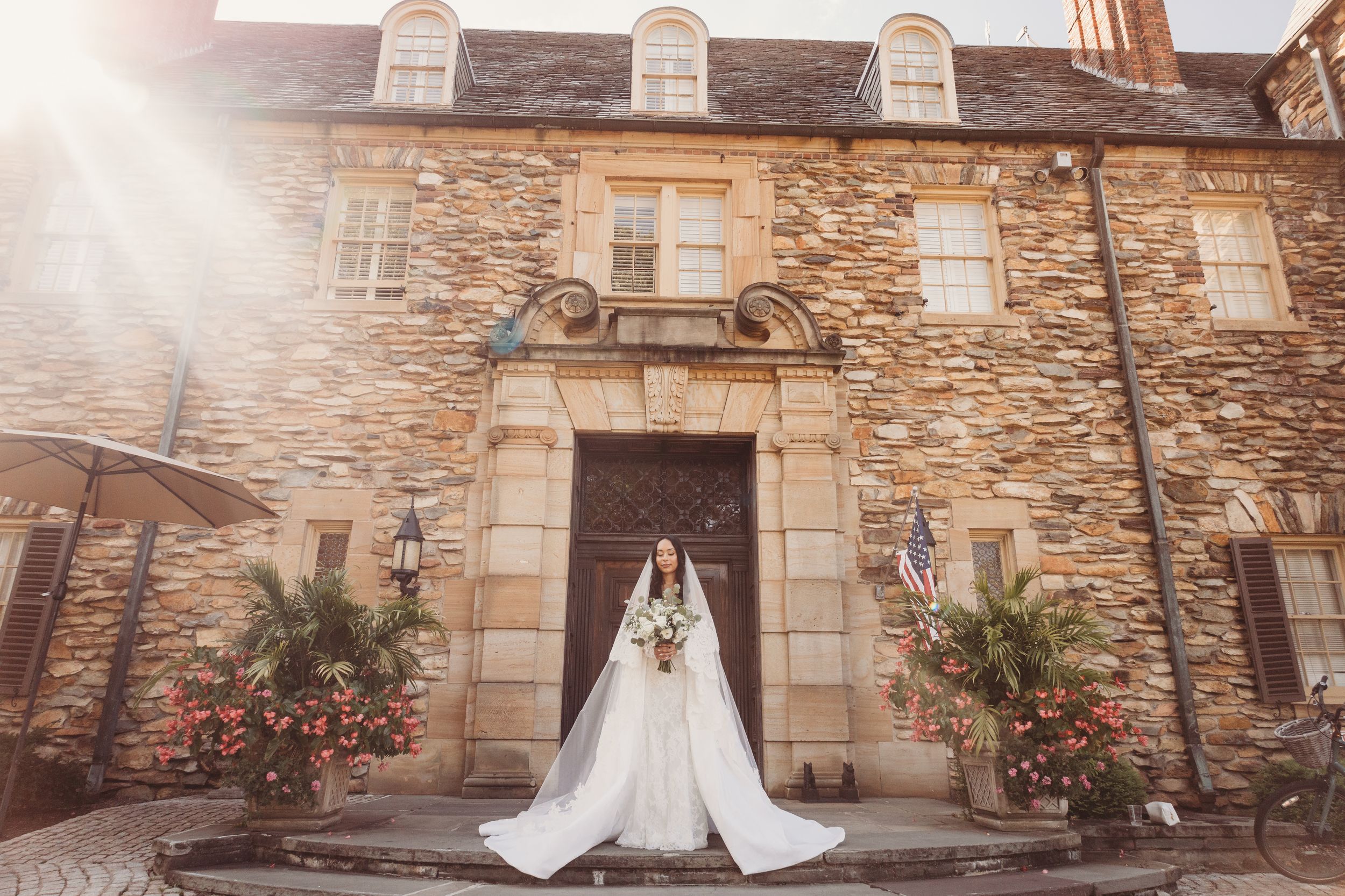 Bride in white gown and veil stands before a stone building entrance with floral arrangements in sunlight.