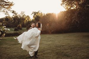 Groom carrying bride in white dress at golden hour sunset in lush green garden during outdoor wedding celebration.