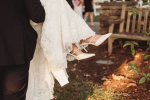 Bride in lace wedding dress being carried by groom, showing white strappy heels against garden backdrop.