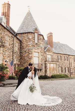 Bride and groom share a romantic dip kiss on cobblestone outside a grand stone castle wedding venue.