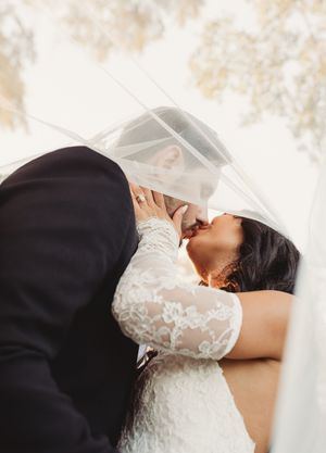 Bride and groom share a passionate kiss under a flowing veil in a dreamy outdoor wedding portrait.