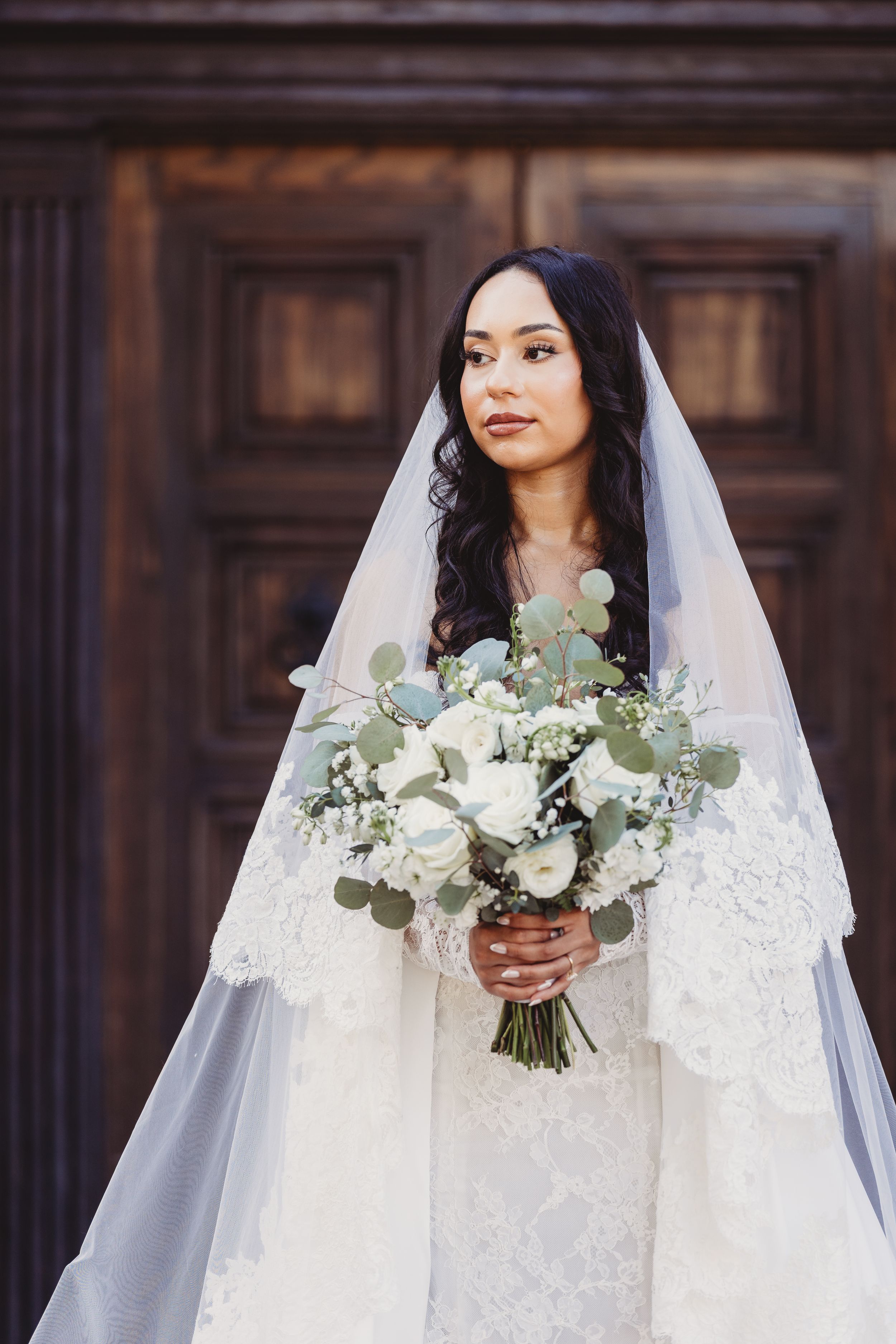 Bride in lace wedding dress and veil holds white rose bouquet before ornate wooden doors.