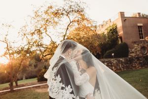 Couple kisses under flowing lace veil at golden sunset beside historic stone building.