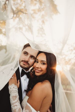 Smiling bride and groom share romantic moment under lace veil in warm golden sunlight.
