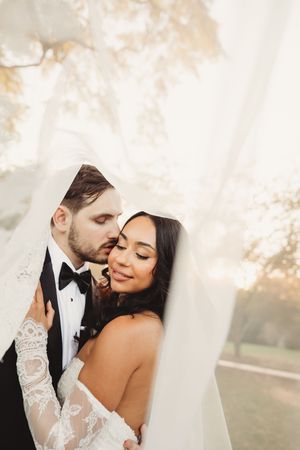 Groom tenderly kisses bride on cheek as her veil wraps around them in a dreamy golden light portrait.
