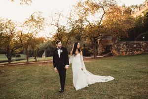 Bride and groom walk hand in hand through golden autumn garden with stone wall backdrop at sunset.