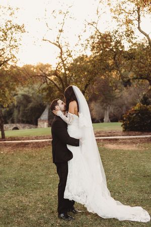 Groom embraces bride in lace wedding gown and veil during golden hour in lush outdoor garden.