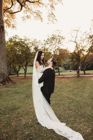 Groom lifting bride in white gown and veil outdoors at golden hour among trees on lush green grass.