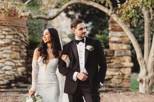 Bride in lace gown and groom in black tuxedo walk arm in arm outdoors near stone arch.