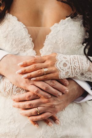 Close-up of bride and groom hands showing gold wedding rings on lace wedding gown.