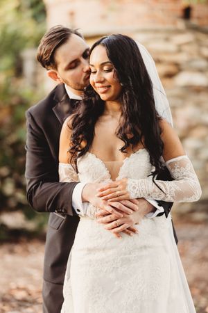 Groom embraces bride from behind near rustic stone wall as she smiles in a warm outdoor wedding portrait.