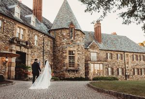 Bride and groom walk hand in hand toward elegant stone manor on cobblestone path.