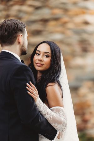 A bride in a lace gown and veil gazes lovingly at her groom in a black suit during an outdoor wedding ceremony.