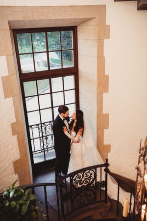 Bride and groom share a romantic moment on elegant spiral staircase by tall window in a classic venue.