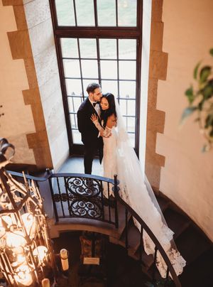 Bride and groom embrace on elegant spiral staircase by tall window, bride in lace gown with veil, groom in black tuxedo.