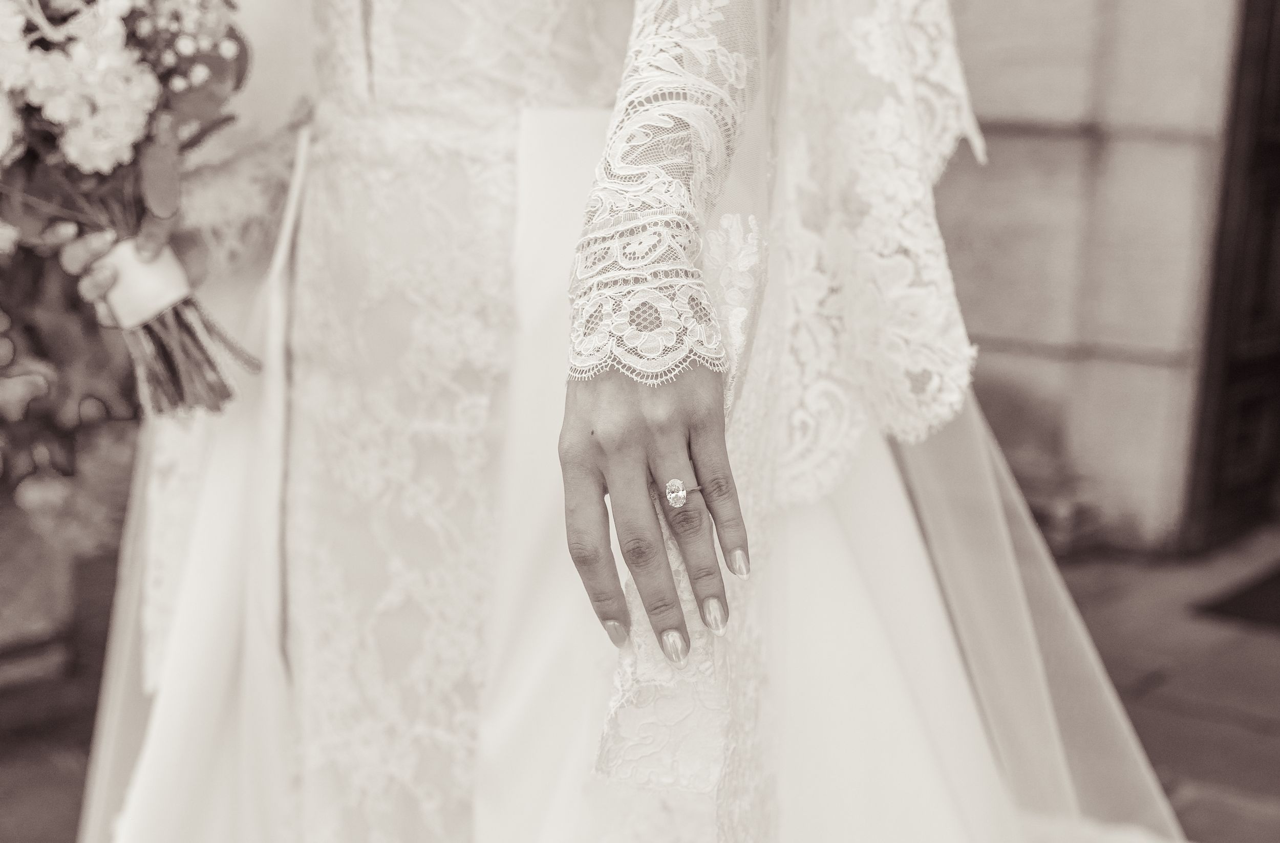 Bride in lace long-sleeve wedding dress displaying diamond engagement ring, holding bouquet, black and white photo.