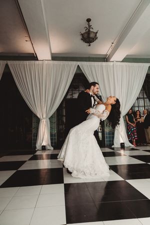 Bride and groom share romantic first dance dip on black and white checkered floor with white drape backdrop.