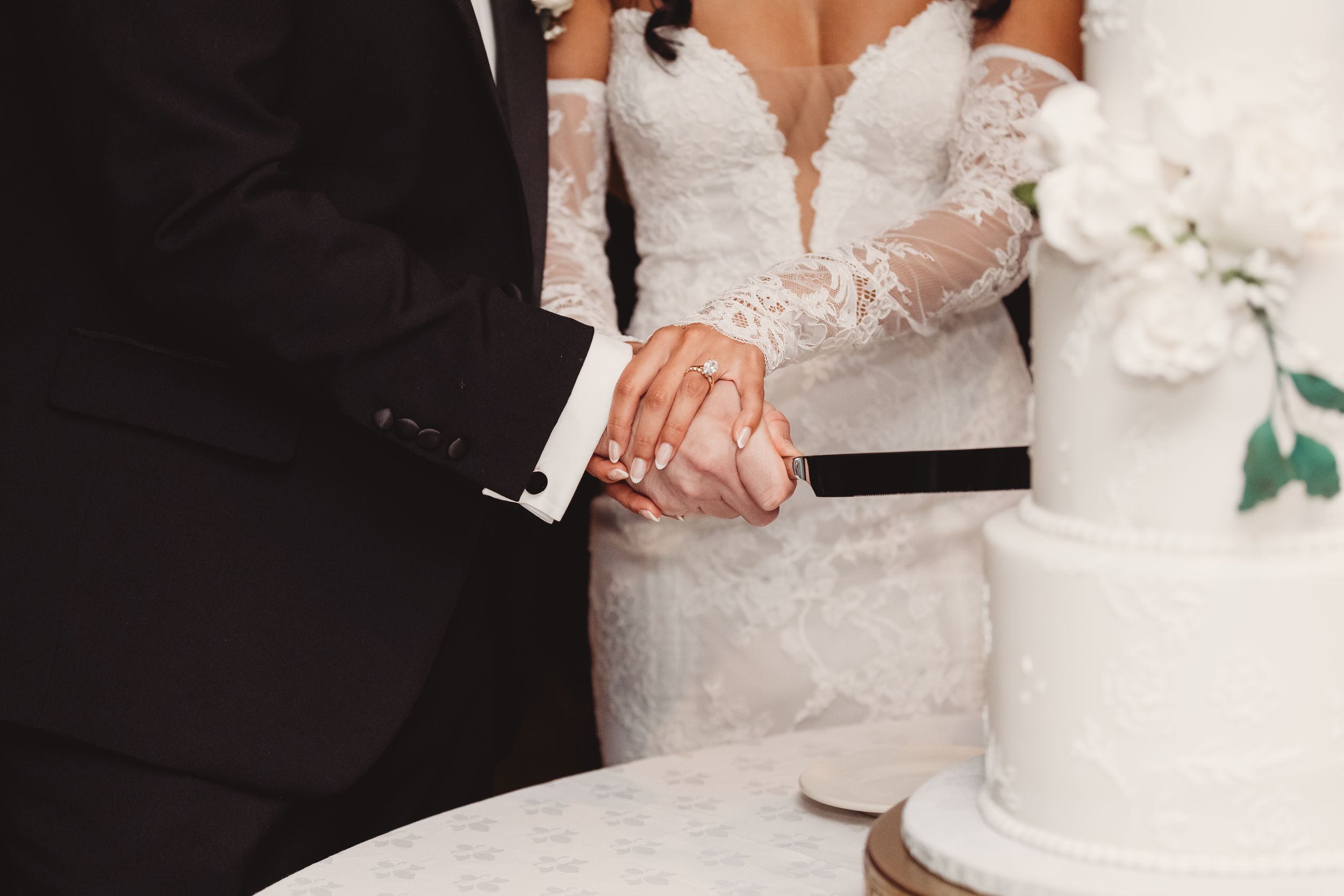Bride and groom cutting white wedding cake together, bride in lace gown, groom in black suit.