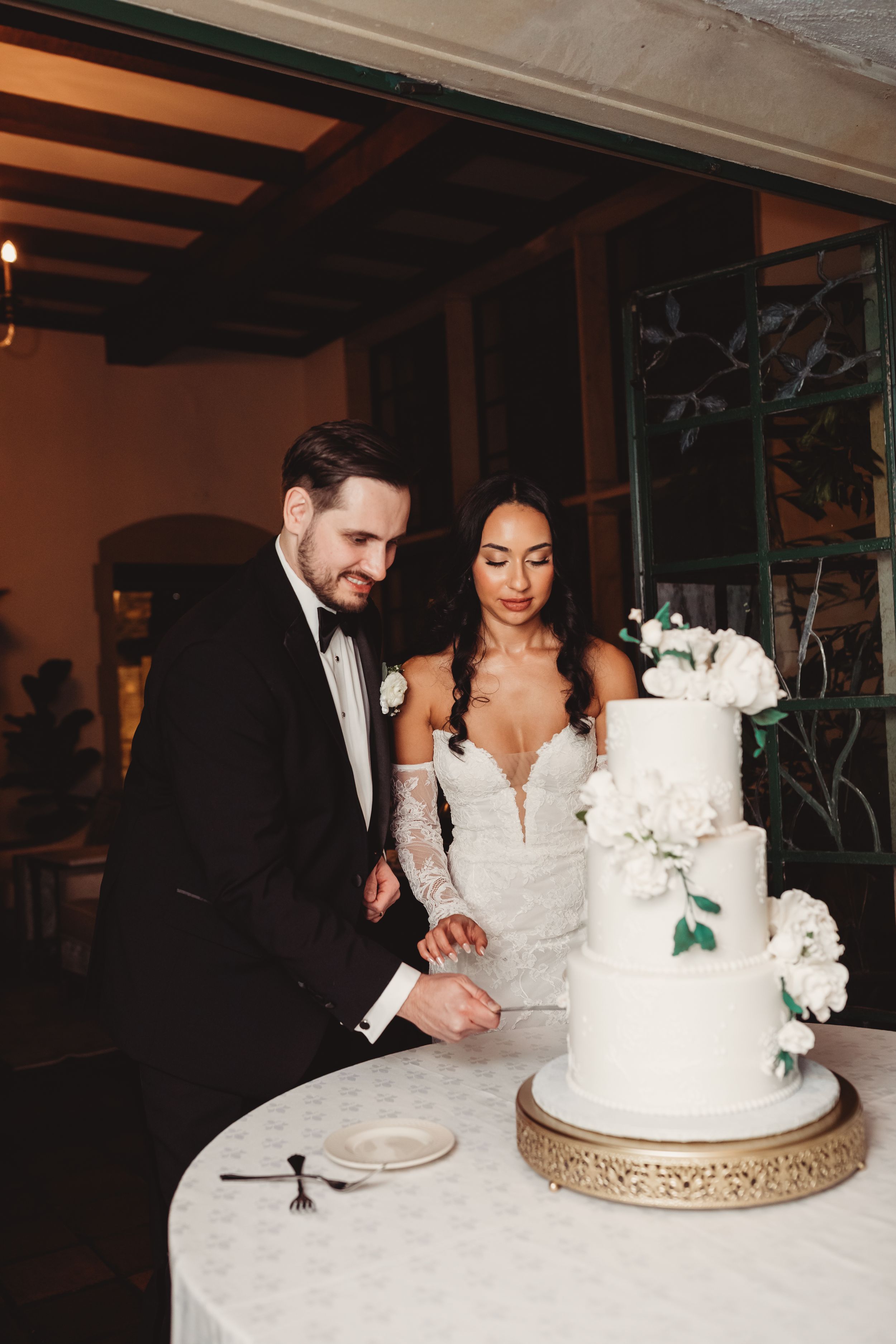 Bride and groom cut elegant three-tier white wedding cake adorned with fresh white flowers and greenery.