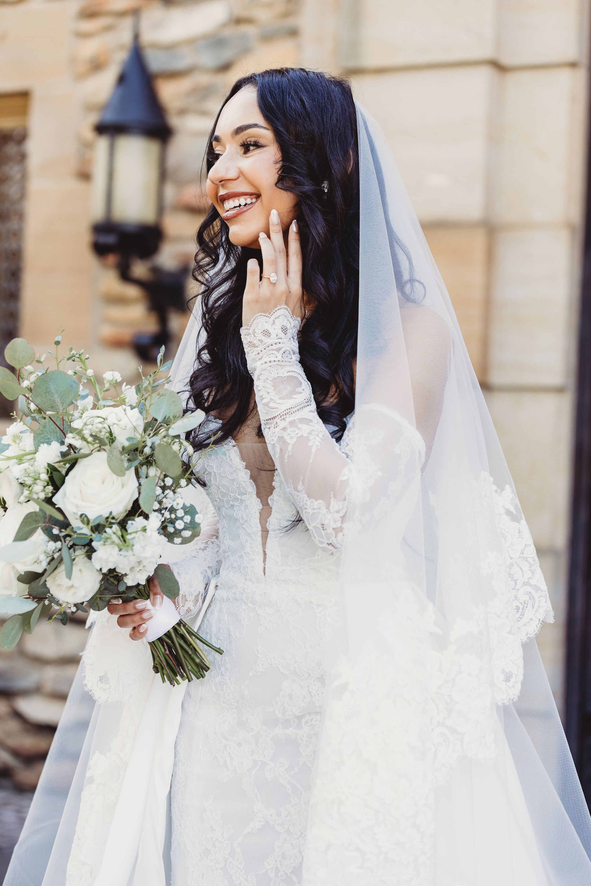 Smiling bride in lace wedding dress with veil holds white floral bouquet outdoors.