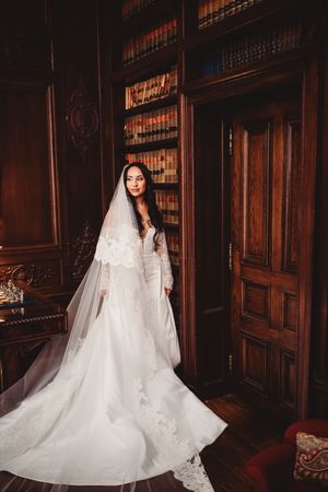 Elegant bride in lace wedding gown and veil poses in a grand library with dark wood bookshelves and ornate door.