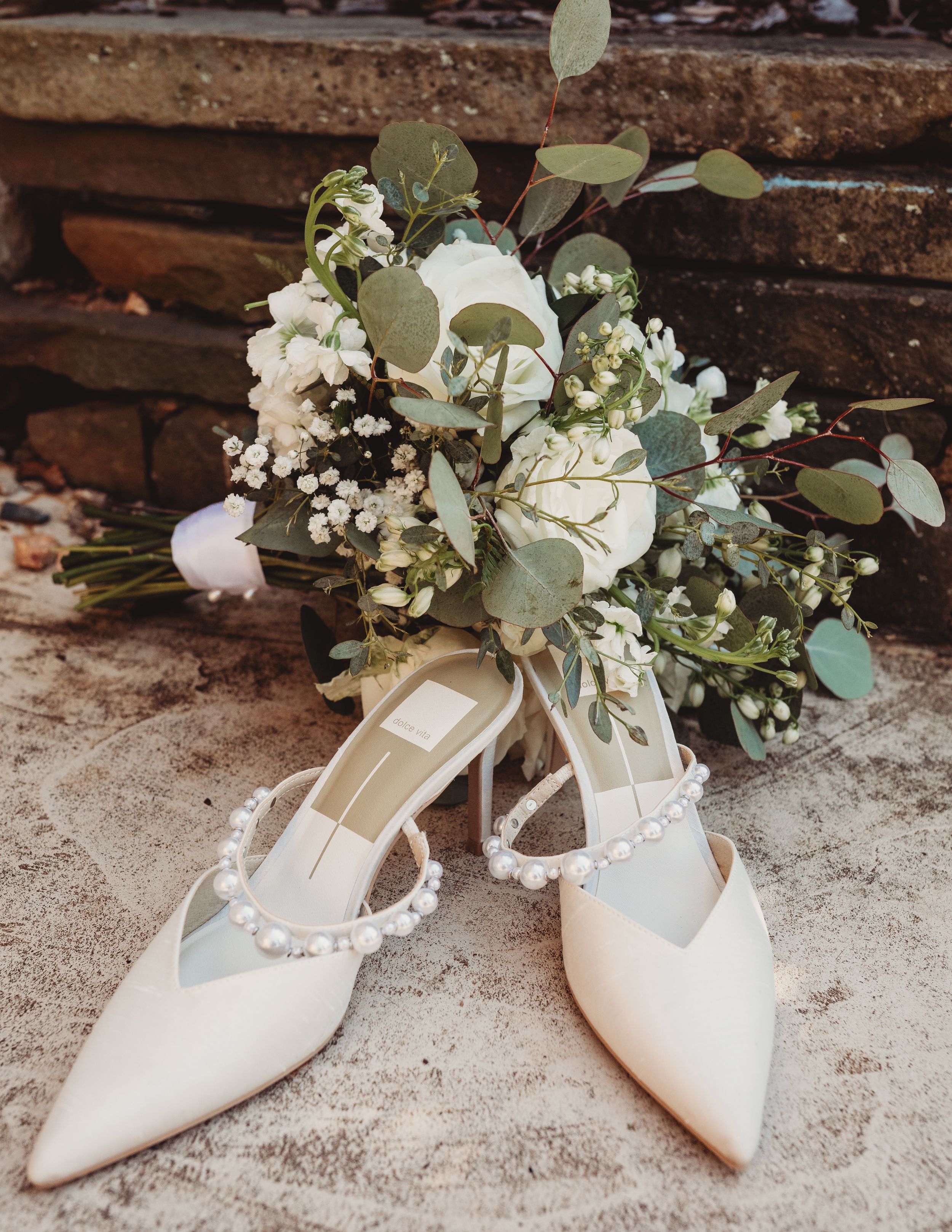Elegant white bridal heels with pearl straps beside a lush eucalyptus and baby's breath wedding bouquet on stone steps.