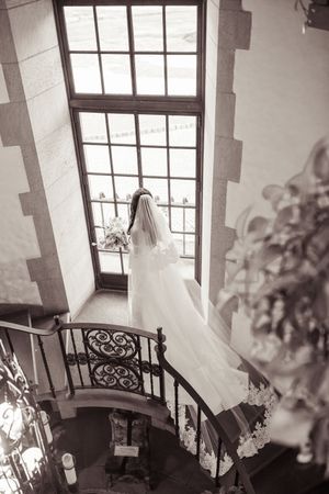 Bride in white gown and veil stands on spiral staircase gazing out tall window in elegant venue.