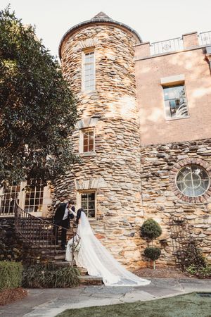 Bride and groom share a romantic moment on stone steps outside a stunning castle-like venue with a circular tower.