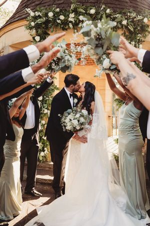 Bride and groom share a kiss under floral arch while wedding party raises hands in celebration.