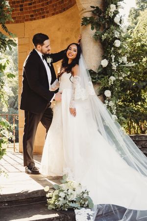 Bride in lace gown and veil with groom in tuxedo posing by floral arch at elegant outdoor wedding ceremony.