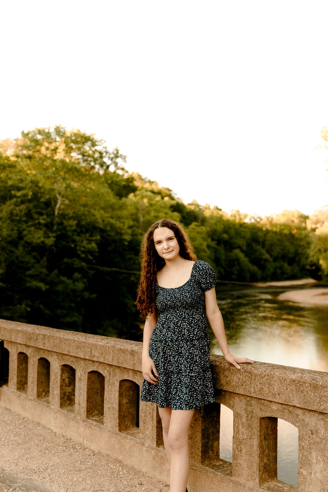 Young woman in floral dress poses on stone bridge with river and trees in golden hour light.