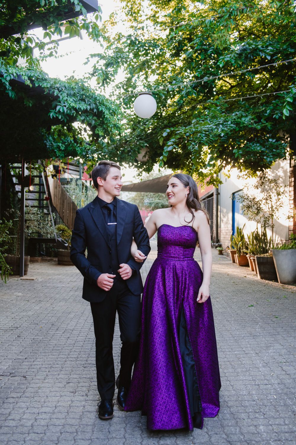 Full-length photo of a matric girl in a purple dress and her date posing at an outdoor and scenic courtyard alleyway.