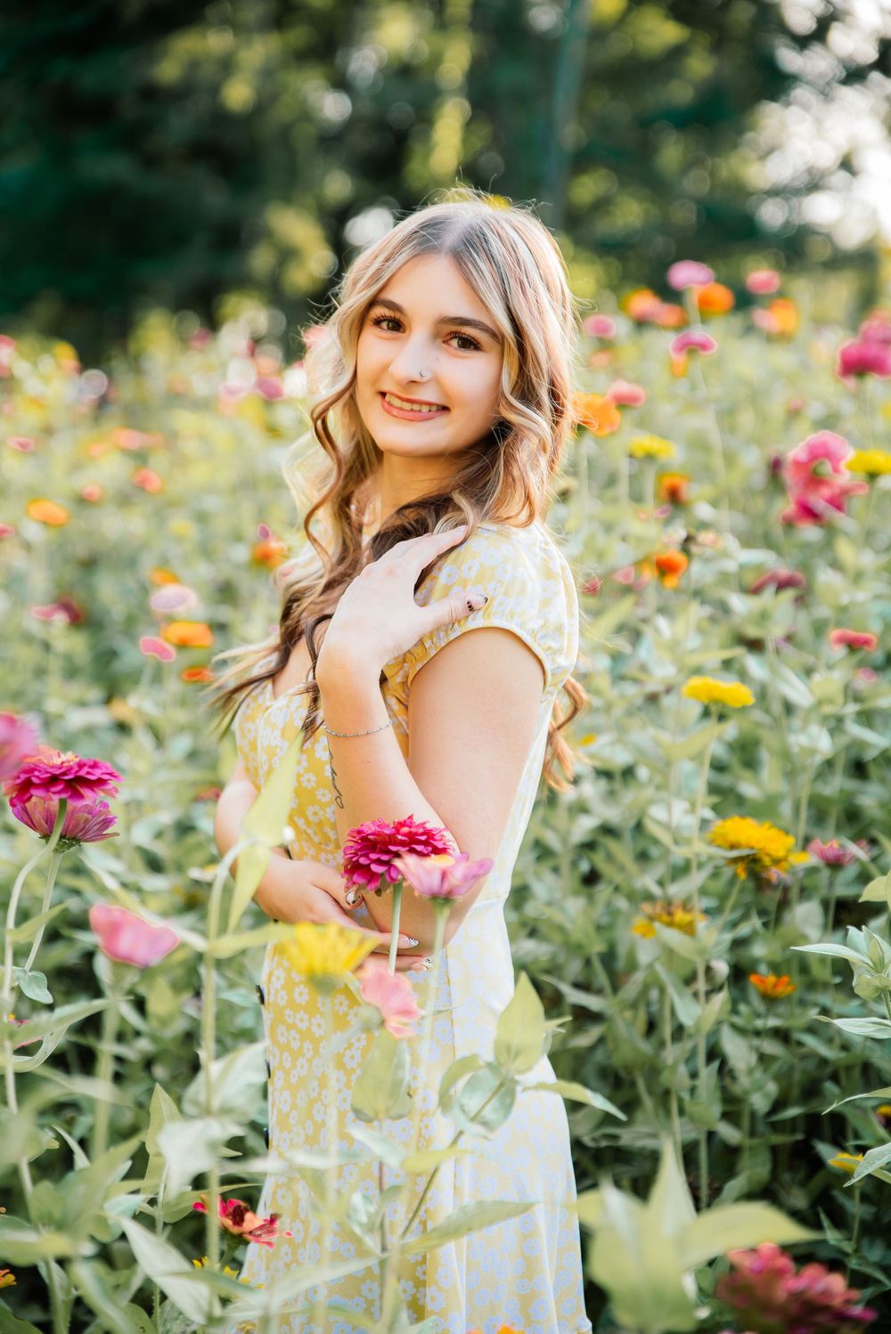 Young woman in yellow floral dress smiling in a vibrant garden field of pink, orange, and yellow zinnia flowers.