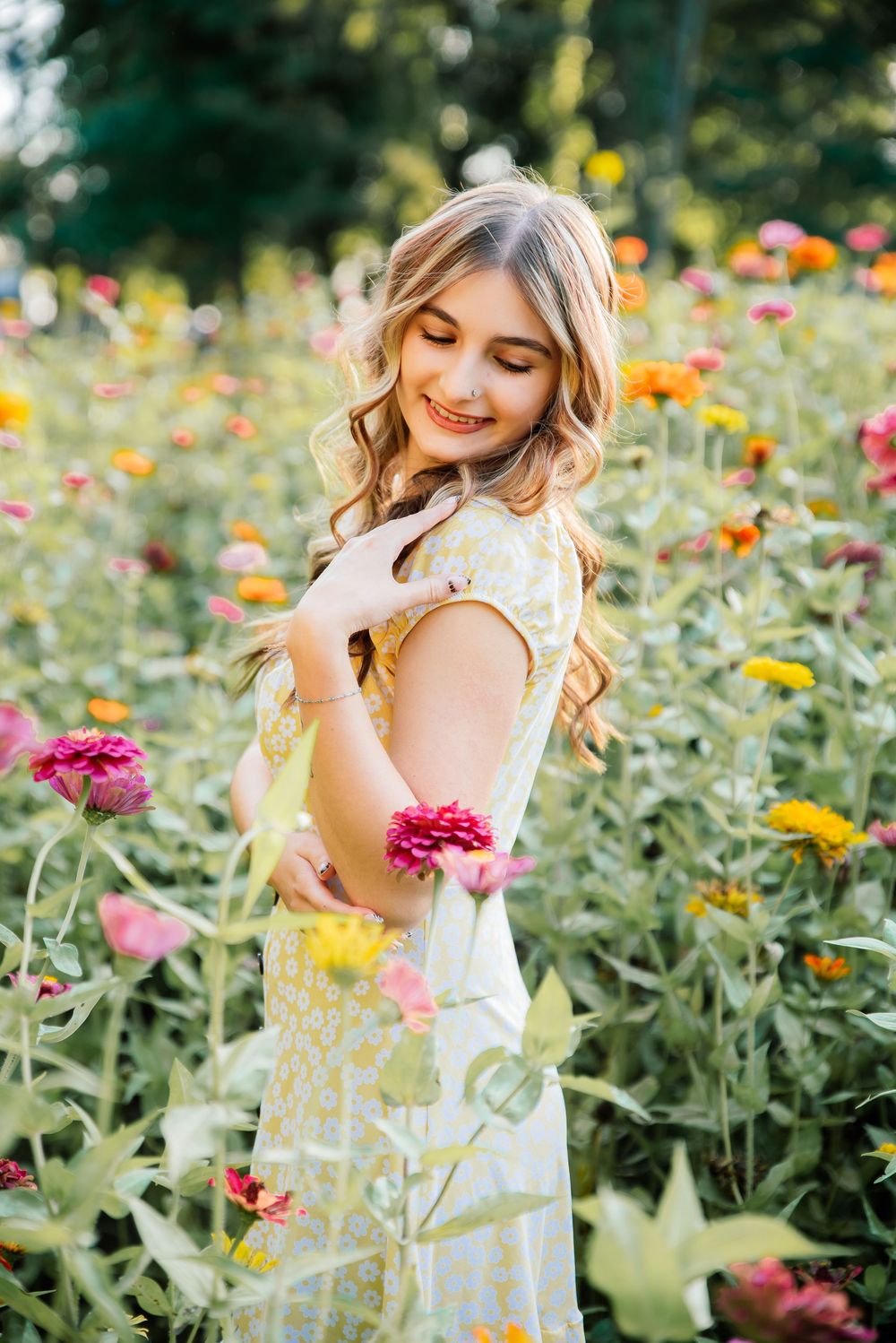 Young woman in yellow floral dress smiling while holding a pink zinnia in a vibrant summer flower field.