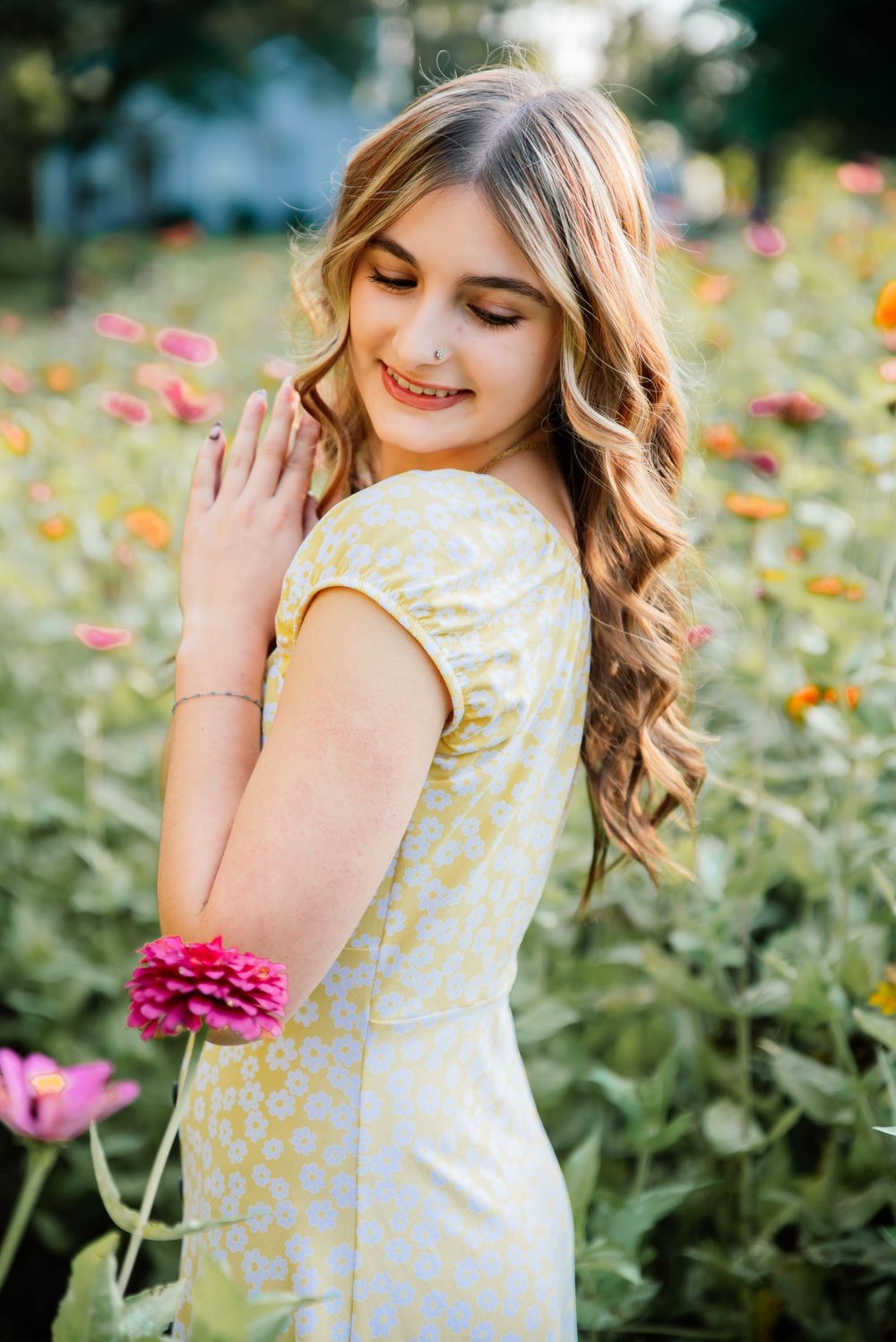Young woman in yellow floral dress smiling among colorful zinnia flowers in a garden.