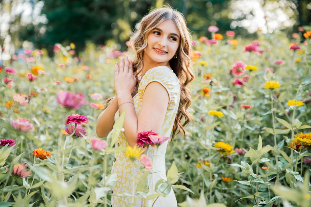Young woman in yellow dress smiling in a vibrant flower field with pink and yellow blooms on a sunny day.