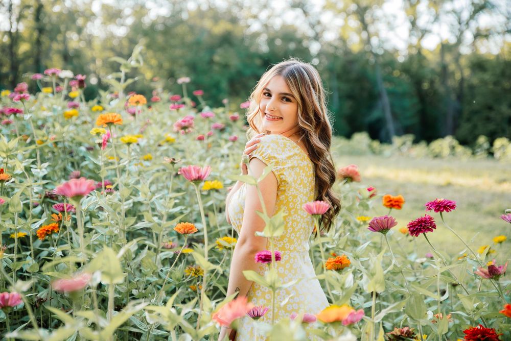 Smiling young woman in yellow floral dress standing among colorful zinnia flowers in a sunny garden.