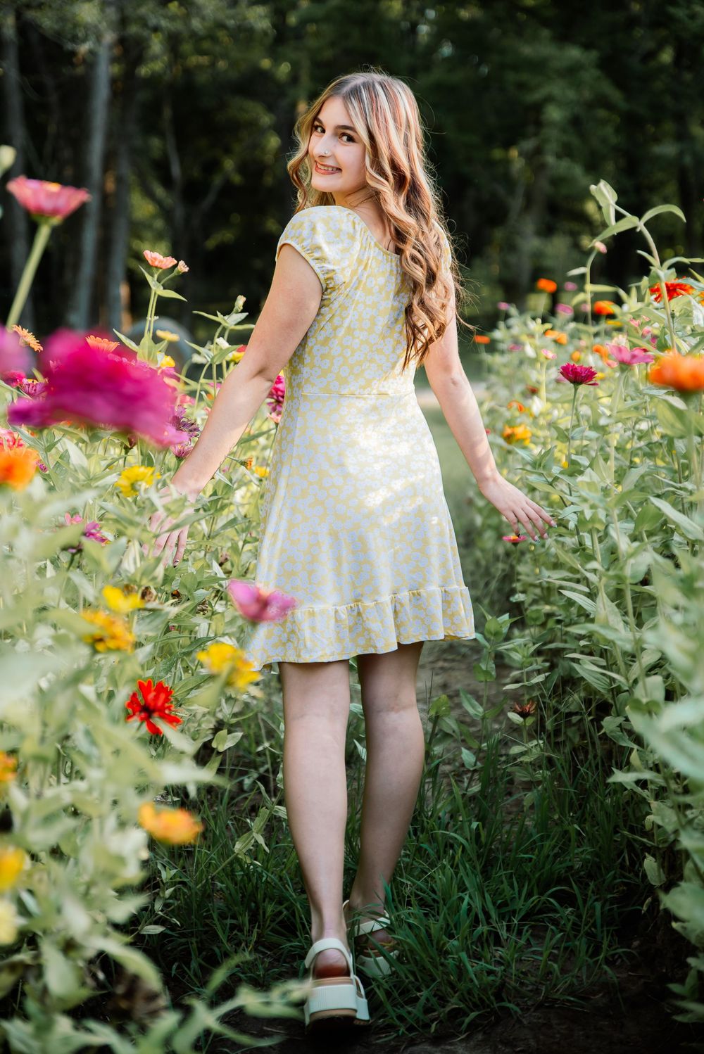 Young woman in yellow floral dress walks through vibrant garden of colorful zinnias, glancing back with a smile.