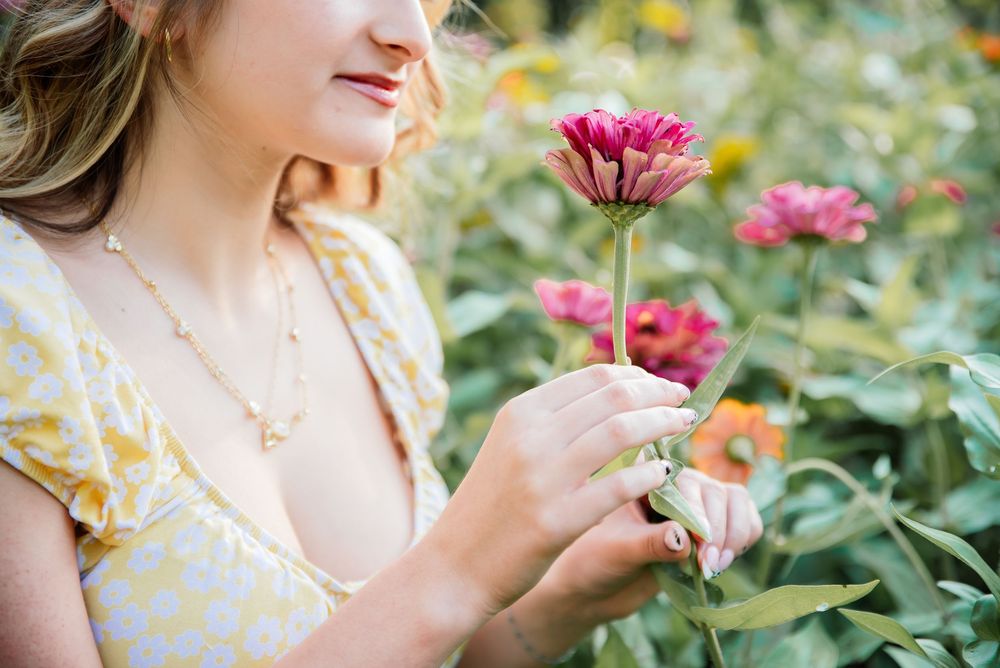 Woman in yellow floral dress holding a pink zinnia flower in a lush garden.