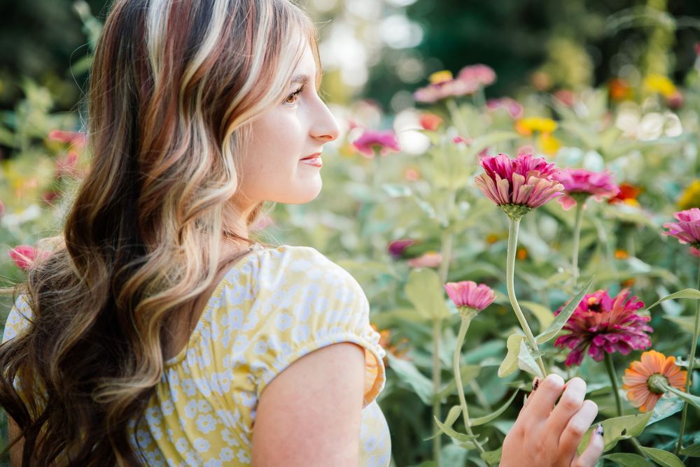 A young woman with wavy hair holds a pink zinnia bloom while standing in a vibrant summer flower garden.