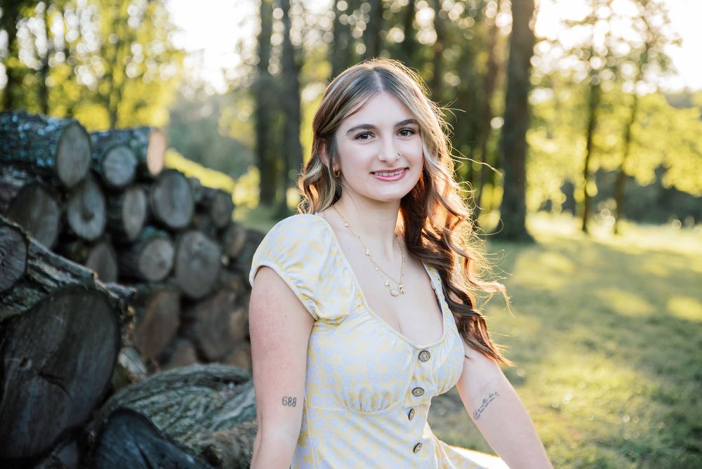 Young woman in yellow dress smiling outdoors near stacked logs in a sunlit forest setting.