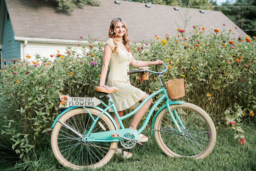 Woman in yellow dress posing with a mint green vintage bicycle adorned with a flower basket in a colorful garden.