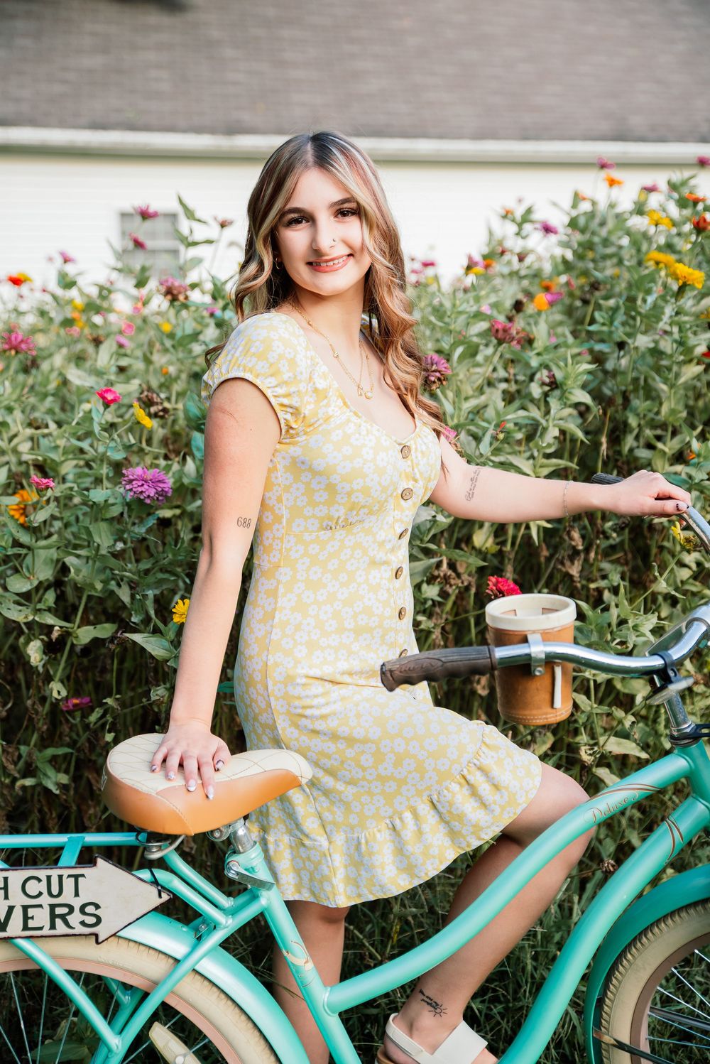 Young woman in yellow floral dress posing with mint green bicycle in front of colorful flower garden.