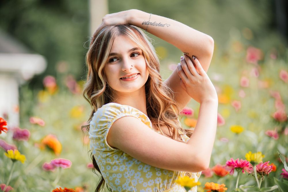 Young woman posing gracefully in a vibrant flower field, smiling softly, wearing a floral dress on a sunny day.