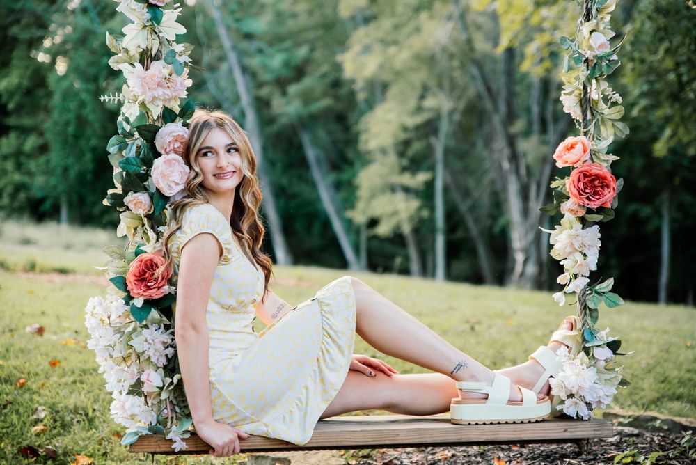 Young woman in yellow dress sitting on floral swing surrounded by pink roses in a lush green outdoor setting.