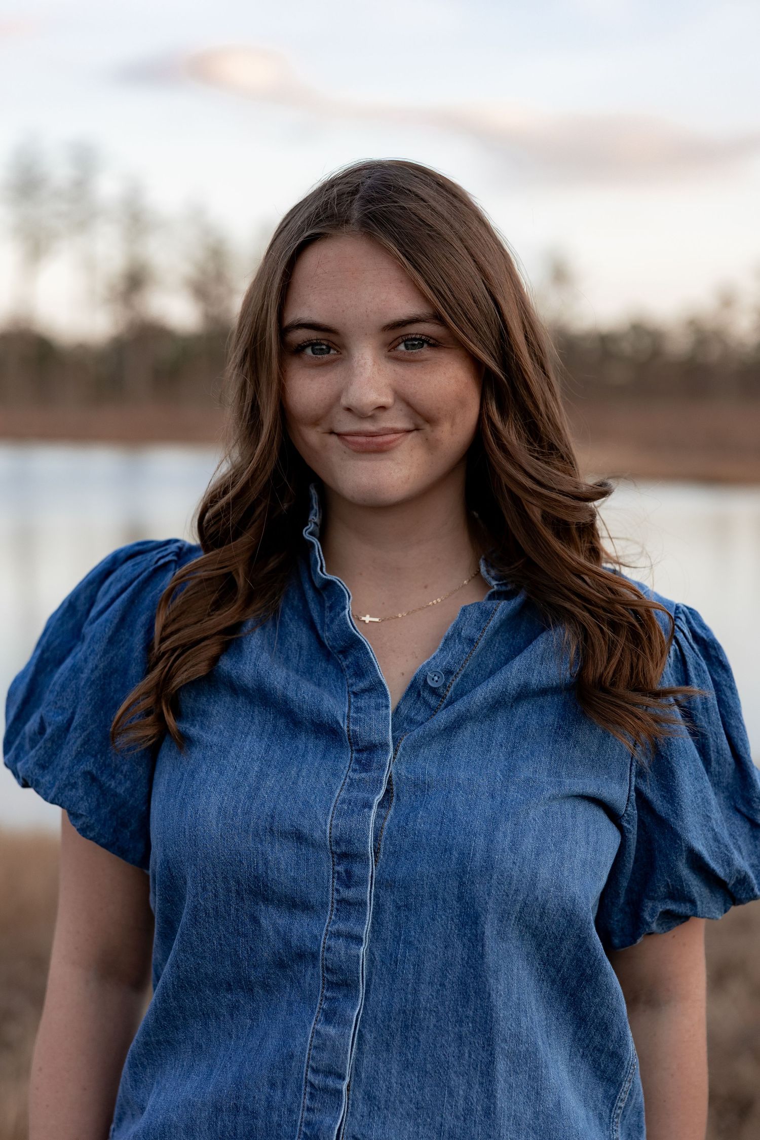 Young woman with brown hair smiling outdoors in a denim shirt near a lake at sunset.