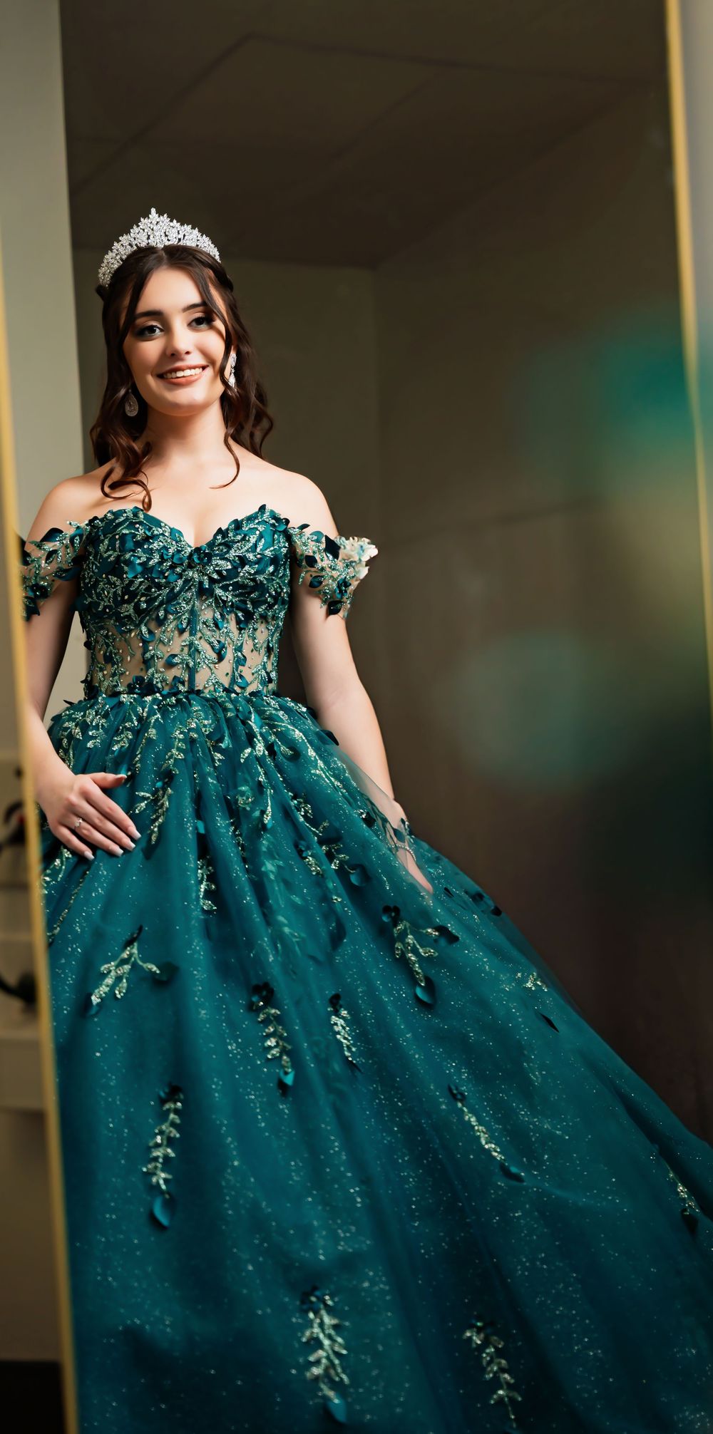 Young woman in a teal off-shoulder ball gown with floral appliques and silver tiara, smiling elegantly.