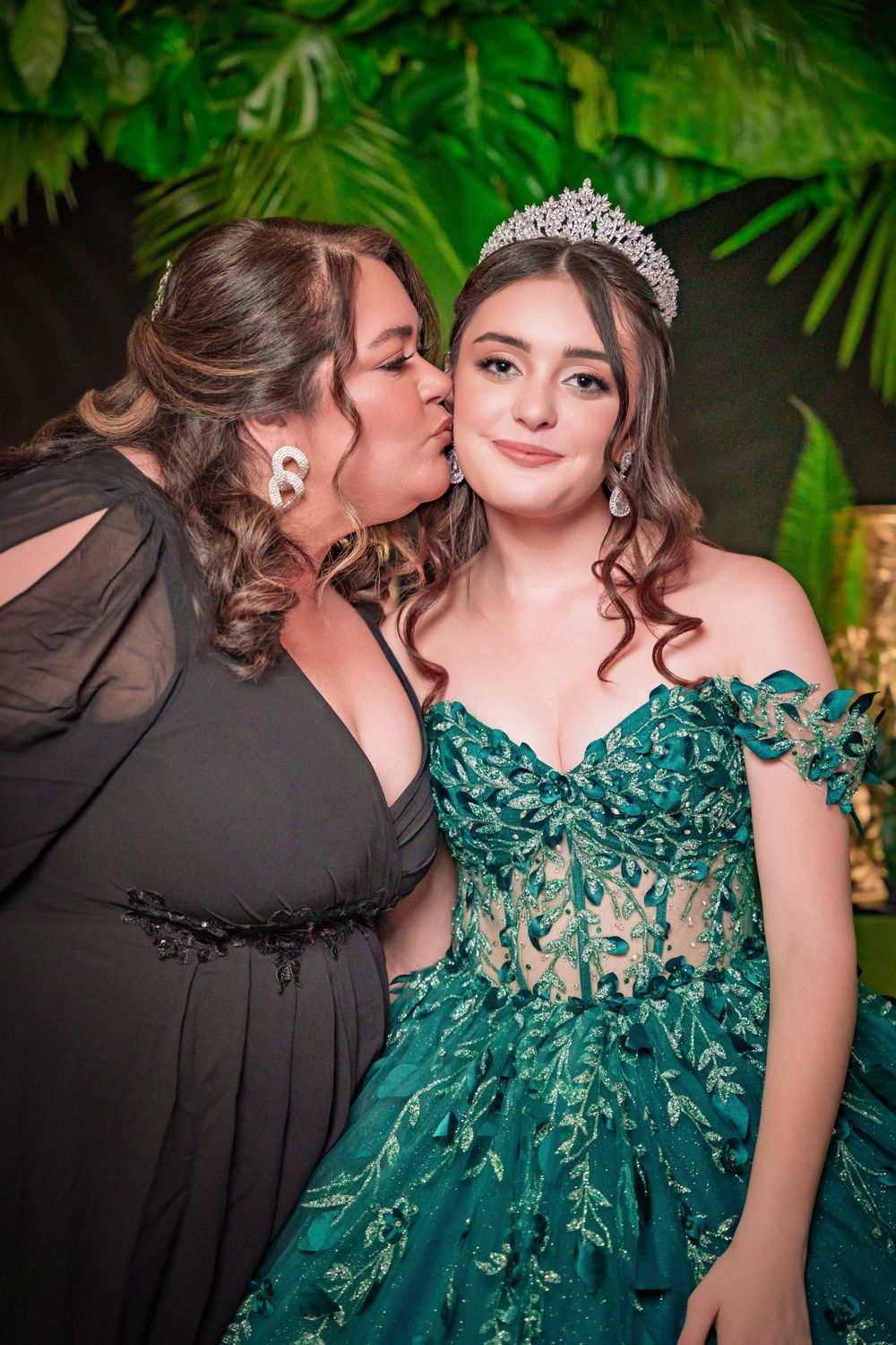 A woman in black dress kisses a young girl wearing a green gown and tiara at a quinceañera celebration.
