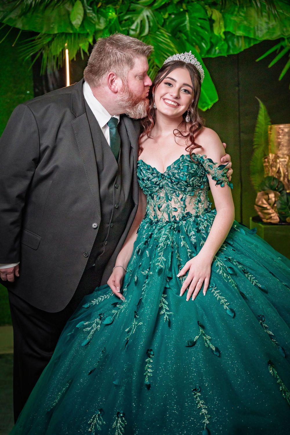 Man in suit kisses girl wearing green quinceañera ball gown and tiara at elegant celebration.